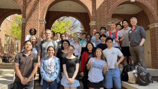 A large, diverse group of students and faculty pose together for a commemorative photo on the steps of a brick building with prominent arched entryways.