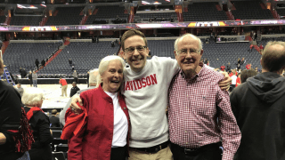 A smiling young man in a white Davidson sweatshirt stands with his arms around an older woman in a red jacket and an older man in a checkered shirt in a basketball arena.