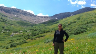 A smiling woman stands in a lush, green mountain valley filled with yellow and red wildflowers under a blue sky with light clouds.