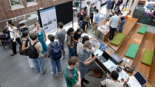 An overhead view of a busy atrium where students and faculty gather around research posters and technical demonstrations during a symposium.