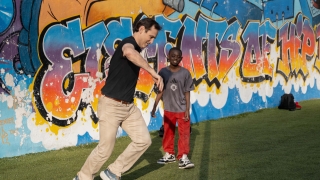 An adult man plays soccer with a young boy in front of a colorful graffiti mural