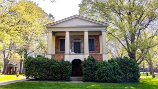 A person sits reading on the upper portico of a historic brick building with classical white columns, surrounded by leafy green trees and hedges on a sunny campus.