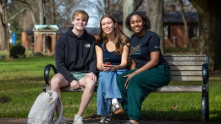 Three students sitting together on a wooden park bench, smiling at the camera.