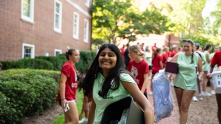 A smiling student in a light green top carries a black storage bin filled with supplies along a brick walkway, surrounded by other students carrying move-in belongings in front of a brick dorm building.