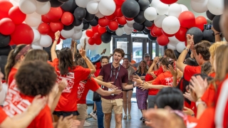 A young man walking down a hallway filled with red, white, and black balloons while crowd members in matching red shirts cheer and high-five him.