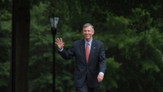 A man in a dark suit, light blue shirt, and red patterned tie smiles and waves while walking down a brick pathway lined with trees and lampposts.