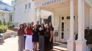A diverse group of nine students smiling together for a group photo under the columned portico of a white "Admission" building.
