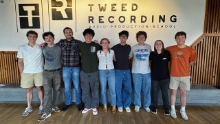 A group of eight young adults stands in a line with their arms around each other's shoulders, posing for a photo inside a modern room in front of a wall with a logo that reads "Tweed Recording Audio Production School."