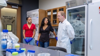 Dr. Thurtle-Schmidt, wearing a white lab coat, speaks with two student researchers wearing protective blue gloves inside a science laboratory.