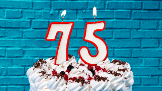 A cake in the foreground, holding candles in the shape of "75", against a blue brick wall of a retro soda shop