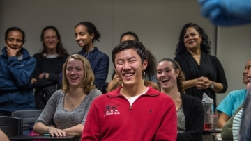 Students are framed through blue gloves of speaker, laughing, with Nail Mamoon in the background