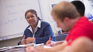 Professor Brenda Flanagan sits with students in class and gives lecture while students take notes