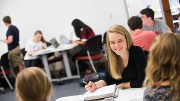 Student talks to classmates sitting at same table. Rest of class is out of focus in the background