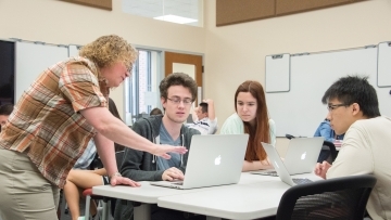 Professor Heyer stands near table and talks to three students who are using laptops in her class