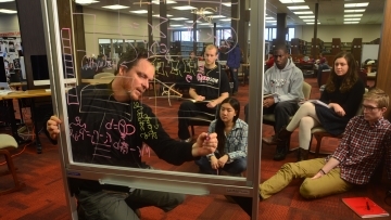 Professor Tim Chartier writes on a glass white board while leading a class to a group of students in the library