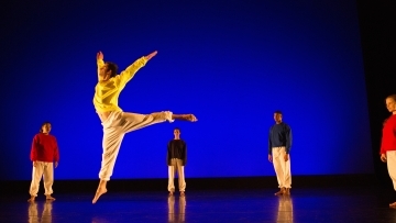 Student leaps into the air in front of blue lights while other dancers on stage stand still