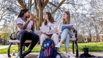 Three students sit on a picnic bench and converse over books