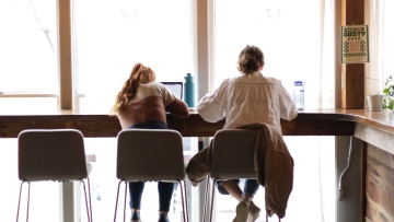 two students sit on high top stools while working on laptops by a window