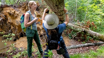 two young women measure a downed tree in the woods while wearing hiking clothes