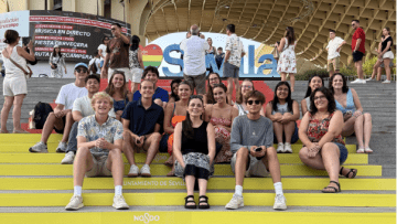 A group of approximately fifteen students sits on yellow and green steps branded with "Ayuntamiento de Sevilla" and "NO8DO" logos, positioned in front of the Setas de Sevilla wooden structure and a colorful "Sevilla" sign.