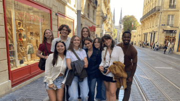 A diverse group of nine smiling students poses on a cobblestone street in Bordeaux next to an "Altermundi" storefront, with tram tracks and distant cathedral spires visible in the background.