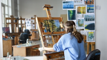An art student in a light blue sweatshirt sits at a wooden easel painting a landscape, with their workspace and various reference images reflected in a large studio mirror.