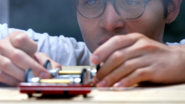 A close-up, eye-level shot shows a student wearing glasses leaning in with intense focus to adjust the wheels of a small model car on a wooden surface.