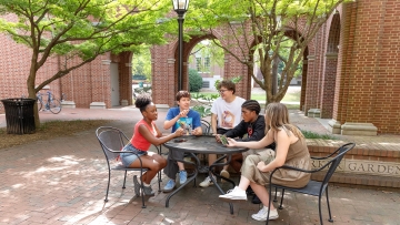 Five students of diverse backgrounds sit around a black metal mesh table in a brick courtyard, engaged in conversation under the shade of green trees and near a brick building with arched walkways.