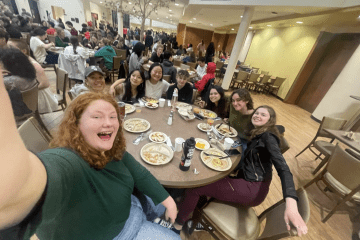 A group of students take a selfie together while sitting around a round dining table in a dining hall