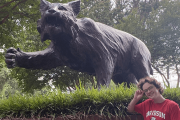a young student wears a Davidson t-shirt while smiling and standing in front of a wildcat statue
