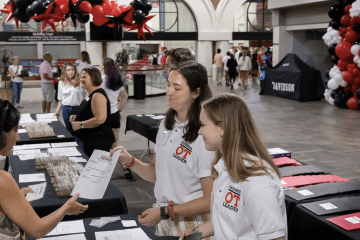 two young women in white polos talk to an older woman around a table with balloons in the background