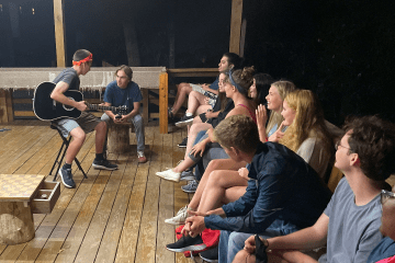 a group of students sit around a cabin as one plays the guitar
