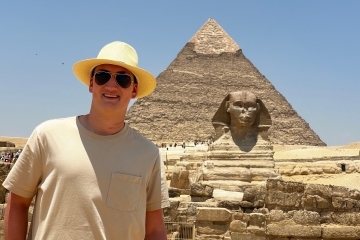 a young white male wears a straw hat while standing in front of an Egyptian pyramid and sphinx