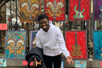 a young Black man wearing a white sweatshirt stands in front of a wall of paintings of musical instruments