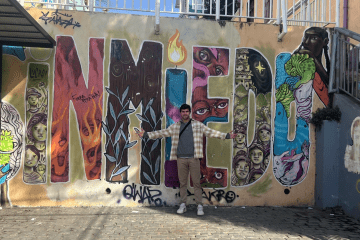a young white man stands in front of a wall that reads " sin miedo"