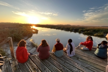 a group of students sit on a dock overlooking marsh at sunset