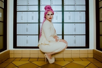 a young white woman in drag crouches in front of a wall of post office boxes