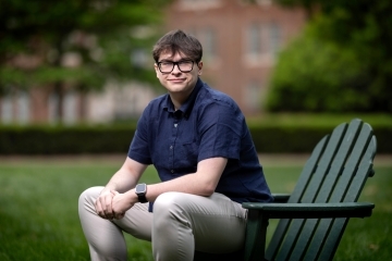 a young white man with a collared blue shirt sitting on a green adirondack chair on a college campus