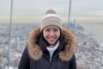a young woman stands on the roof of a skyscraper in New York City, she wears a beanie and big coat and the sky appears to be grey and cloudy