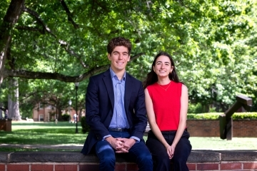 a young man and a young woman sit together on a brick wall in front of green trees