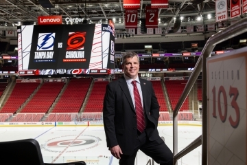 a middle aged white man wearing a black suit with a red tie in front of the Carolina Hurricanes hockey rink