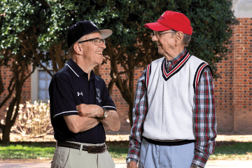 two older men stand together smiling looking at each other in front of a stadium, wearing Davidson College gear