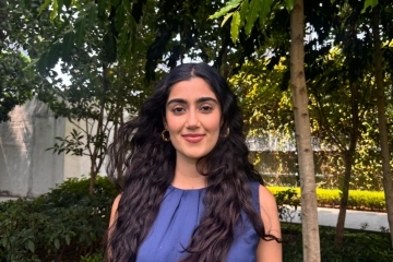 a young woman with long dark curly hair wearing a blue top standing outside in front of a set of green trees