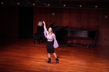 a young woman sings on stage in a purple blouse and black skirt