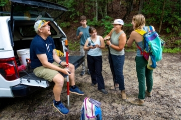 a group of students with their professor gathered around a truck in the woods wearing hiking gear