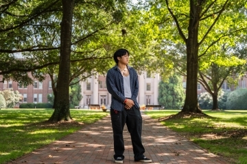 a young man stands on a college campus looking to his side in front of a large building surrounded by trees