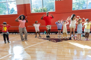 A group of diverse children and young adult leaders stand in a row on a gymnasium floor, smiling and flexing their muscles in a strong pose against a red and white wall.