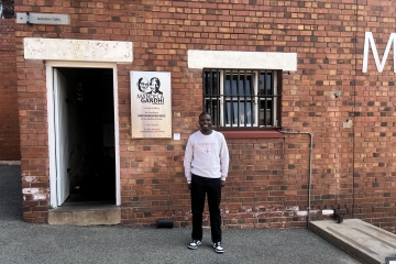 A man in a light grey sweatshirt and black pants stands in front of a red brick building at Constitution Hill in Johannesburg, next to an open doorway labeled "Isolation Cells" and a plaque commemorating the "Mandela Gandhi" exhibition.