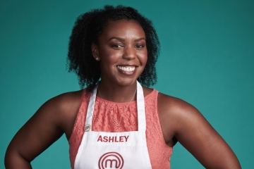 A smiling Black woman with curly hair wearing a white MasterChef apron with the name "ASHLEY" printed on it over a coral lace top, posed against a solid teal background.