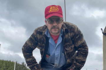 A man with a mustache, sunglasses, and a red Arkansas Razorbacks hat stands outdoors under a cloudy sky wearing a patterned wool jacket over a blue button-down shirt.